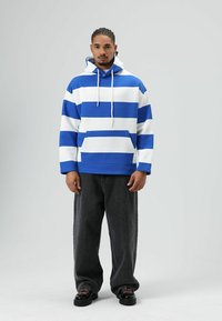 Gestreepte blauwe en witte hoodie met een voorzak, capuchon met trekkoord en lange mouwen, gecombineerd met donkergroene oversized broek en zwarte schoenen.