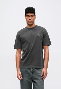 Camiseta de algodón gris con mangas cortas, cuello redondo y sutil logo frontal. Combinada con jeans de mezclilla de lavado claro, el modelo se encuentra de pie contra un fondo plano.