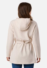 Sweat à capuche beige clair avec une taille cintrée, des manches longues et une texture douce. Détails de couture professionnelle aux bords.