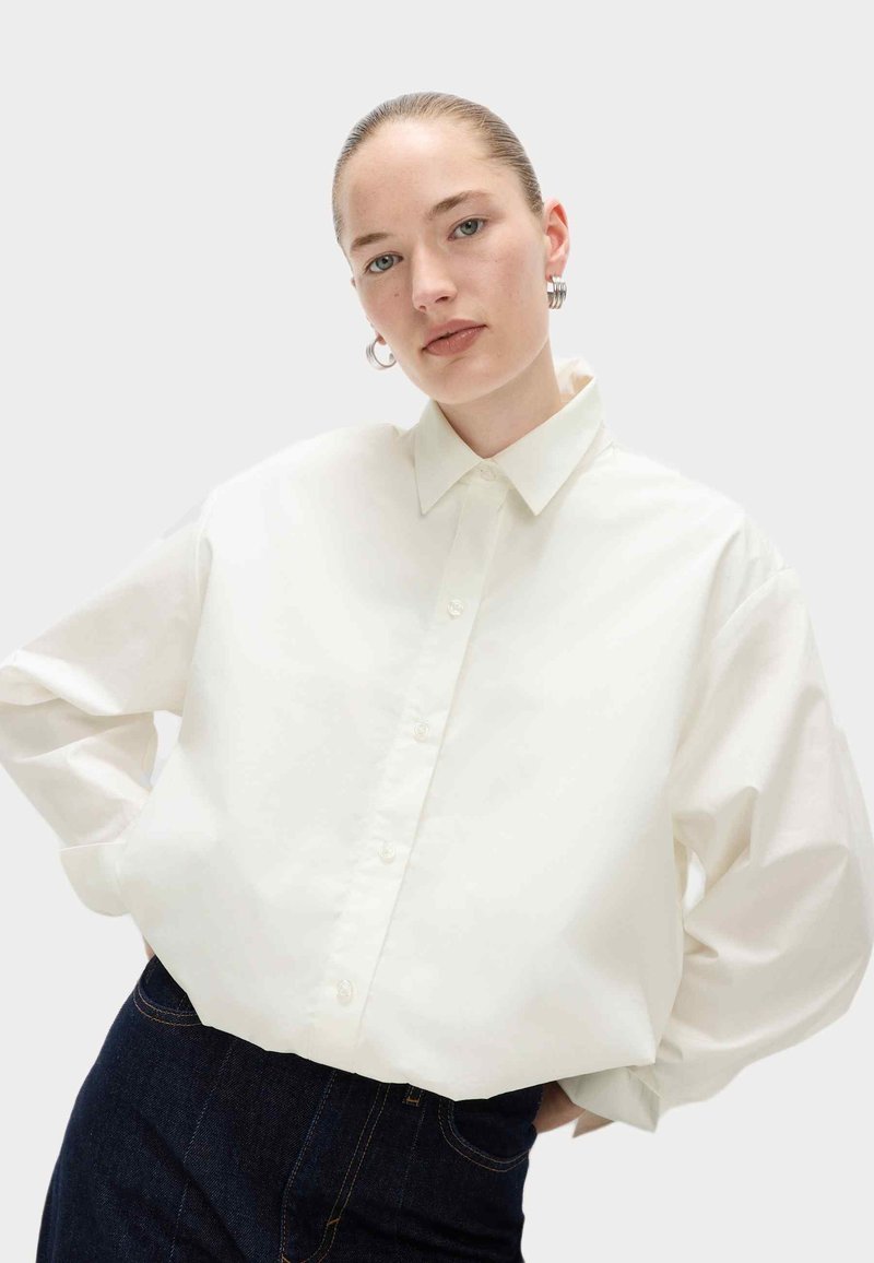 Camicia bianca oversize con colletto a punta, maniche lunghe e una texture liscia, abbinata a jeans in denim scuri.