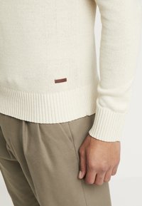 Ljus stickad tröja i krämfärg med ribbade mudd och nederkant, som har en liten brun etikett sydd på sidan. Kombinerad med ljusa byxor.