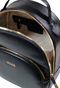 Sort læderrygsæk med lynlås i guldfarve, med et tanfarvet mønstret for, en lille indvendig lomme og præget brandlogo på forsiden.