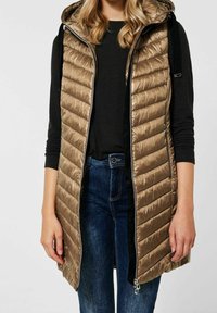 Puffig huva väst i metallisk gyllene tyg med vertikala kanaler, framside dragkedja och sidofickor, kombinerad med en svart topp och jeans.