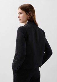 Blazer noir ajusté en tissu texturé, avec des épaules structurées et des boutons sur les manches, offrant une silhouette cintrée et des détails subtils.
