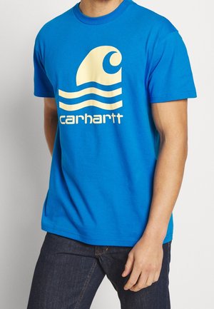 Mężczyzna ubrany w jasnoniebieski t-shirt z krótkim rękawem z beżowym logo fali i tekstem "carhartt", w połączeniu z ciemnymi dżinsami.