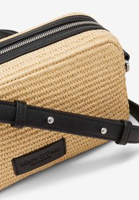 Bolso cruzado de paja tejido en color natural con detalles en cuero negro, que cuenta con un cierre de cremallera y una correa ajustable. Etiqueta de la marca visible.