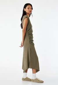 Olivenfarvet midikjole med tekstureret overflade, kapærmer, side-ruche og en asymmetrisk kant. Matchet med beige sneakers og hvide sokker.