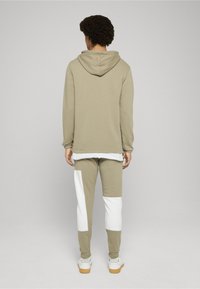 Olivově zelená mikina s kapucí, spárovaná s olivově a bíle barevně blokovanými tepláky. Obsahuje elastické manžety a vrstvený vzhled.