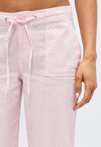 Ροζ λινά παντελόνια με μπροστινή τσέπη και ζώνη με κορδόνι στη μέση, εμφανιζόμενα στη μέση ενός μοντέλου και το δεξί χέρι ακουμπάει δίπλα στο μηρό.