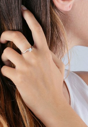 Silberner Ring mit einem zentralen klaren Edelstein, dünner Ringband, getragen an einer Hand, die das Haar berührt, und das Design sowie die Klarheit des Rings zur Schau stellt.