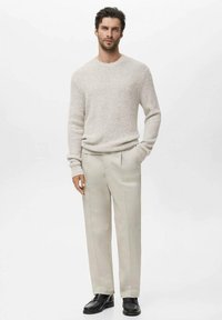 Man staat met één hand in zijn zak, gekleed in een lichtbeige gebreide trui, bijpassende op maat gemaakte broek en zwarte loafers, tegen een effen witte achtergrond.