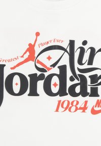 Λευκό μπλουζάκι με μαύρο έντονο κείμενο "Air Jordan" και κόκκινες πινελιές, που περιλαμβάνουν ένα λογότυπο με φιγούρα που πηδάει και το "1984" με κόκκινο.