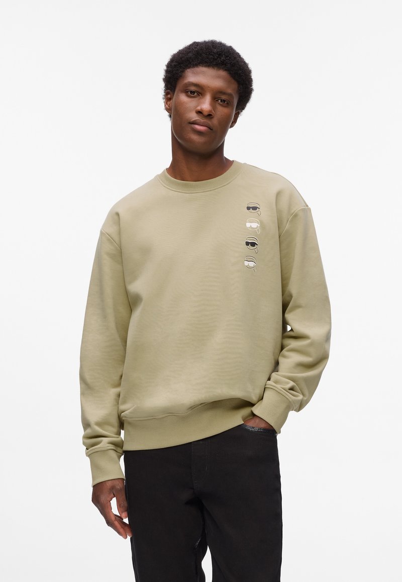 Sudadera de un suave color verde oliva claro, confeccionada en un tejido texturizado. Presenta un escote redondo y un patrón vertical de gafas de sol estilizadas en la parte frontal.