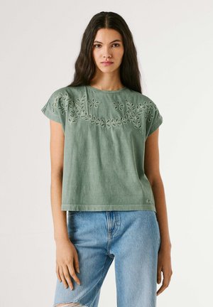 Jonge vrouw met lang donker haar, gekleed in een groene geborduurde top en lichtblauwe spijkerbroek, staand tegen een effen achtergrond.