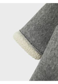 Tissu gris avec une texture douce et duveteuse, présentant un poignet en laine de couleur crème. La couture est soigneusement réalisée, mettant en valeur le contraste des tissus.
