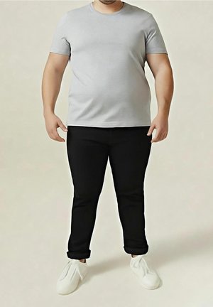 Mand iført en lysegrå kortærmet skjorte, sorte bukser rullet op ved anklerne og hvide slip-on sko, stående mod en ensfarvet baggrund.