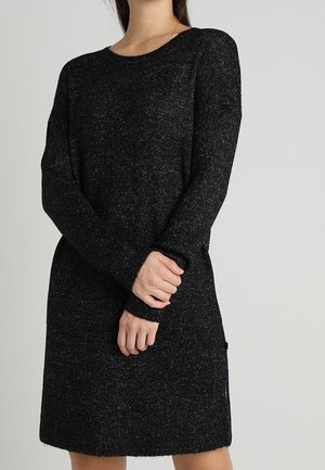 Nainen, joka on pukeutunut pitkähiharaiseen mustaan neulemekkoon, kädet ristissä edessä ja vaaleanharmaa tausta.