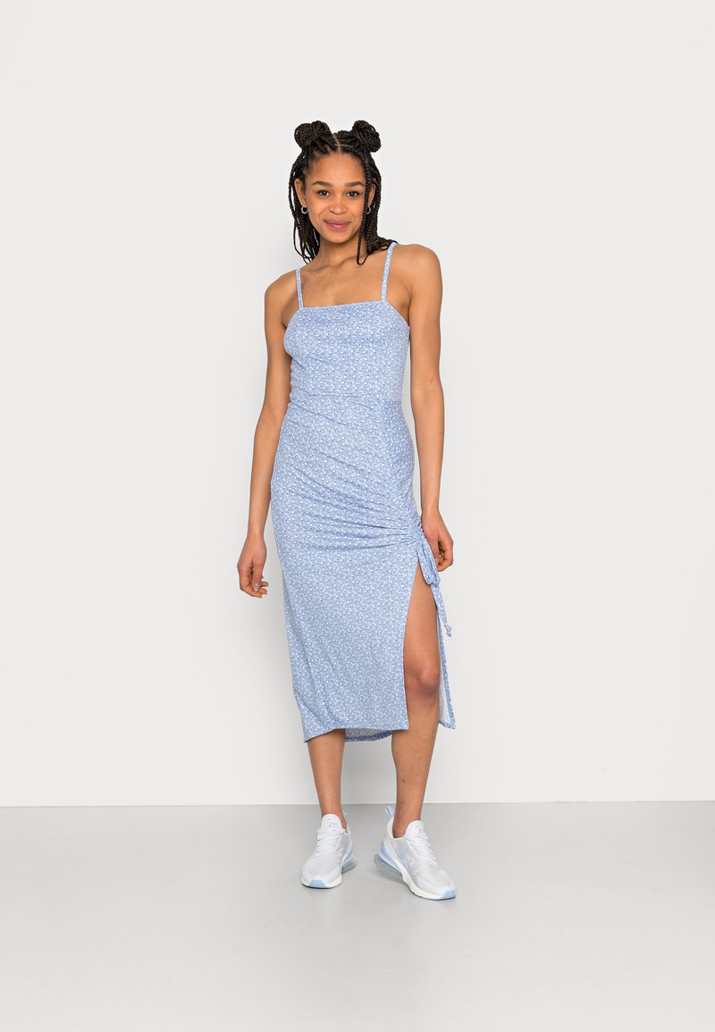 Vestido midi de padrão floral em azul claro, com alças finas ajustáveis, franzido lateral e uma fenda alta, combinado com ténis brancos.
