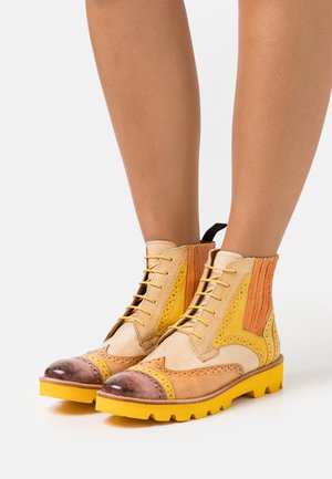 Stivali alla caviglia in pelle beige e marrone con suole gialle. Le caratteristiche includono dettagli brogue, accenti laterali gialli e arancioni, e lacci.