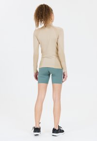 Beige Langarmshirt mit strukturierten Einsätzen, kombiniert mit türkisfarbenen Shorts. Die Person trägt schwarze Sportschuhe. Schlichter Hintergrund.