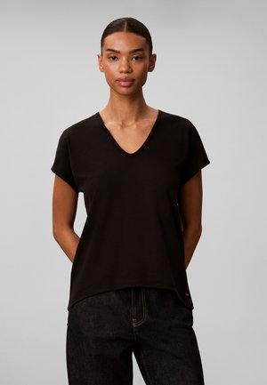 Jeune femme aux cheveux foncés tirés en arrière, portant un t-shirt noir à manches courtes et col en V, ainsi qu'un jean foncé, debout les mains dans le dos.