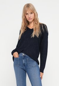 Suéter de malha azul escuro com decote em V e mangas longas, combinado com jeans de cintura alta azul claro com bolsos traseiros. Textura suave.