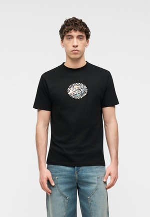 Νέος άνδρας με σγουρά μαλλιά που φοράει μαύρο μπλουζάκι με κυκλικό λογότυπο "Santa Cruz" και ανοιχτόχρωμο μπλε τζιν, στέκεται μπροστά σε απλό φόντο.