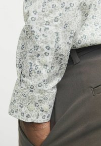 Camisa blanca con un patrón floral y acentos en gris y azul, con puños de botón. Combinada con pantalones oscuros de textura marrón.
