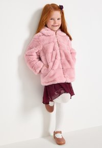 Giacca in finta pelliccia rosa con chiusura a zip e tasche, abbinata a una gonna in pizzo bordeaux e collant bianchi a trama. Scarpe marroni completano l'outfit.