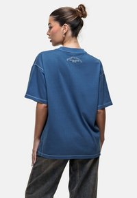 Blauwe oversized katoenen T-shirt met korte mouwen, met een logo op de achterkant. Zachte textuur en casual pasvorm, gecombineerd met donkerdenim broek.