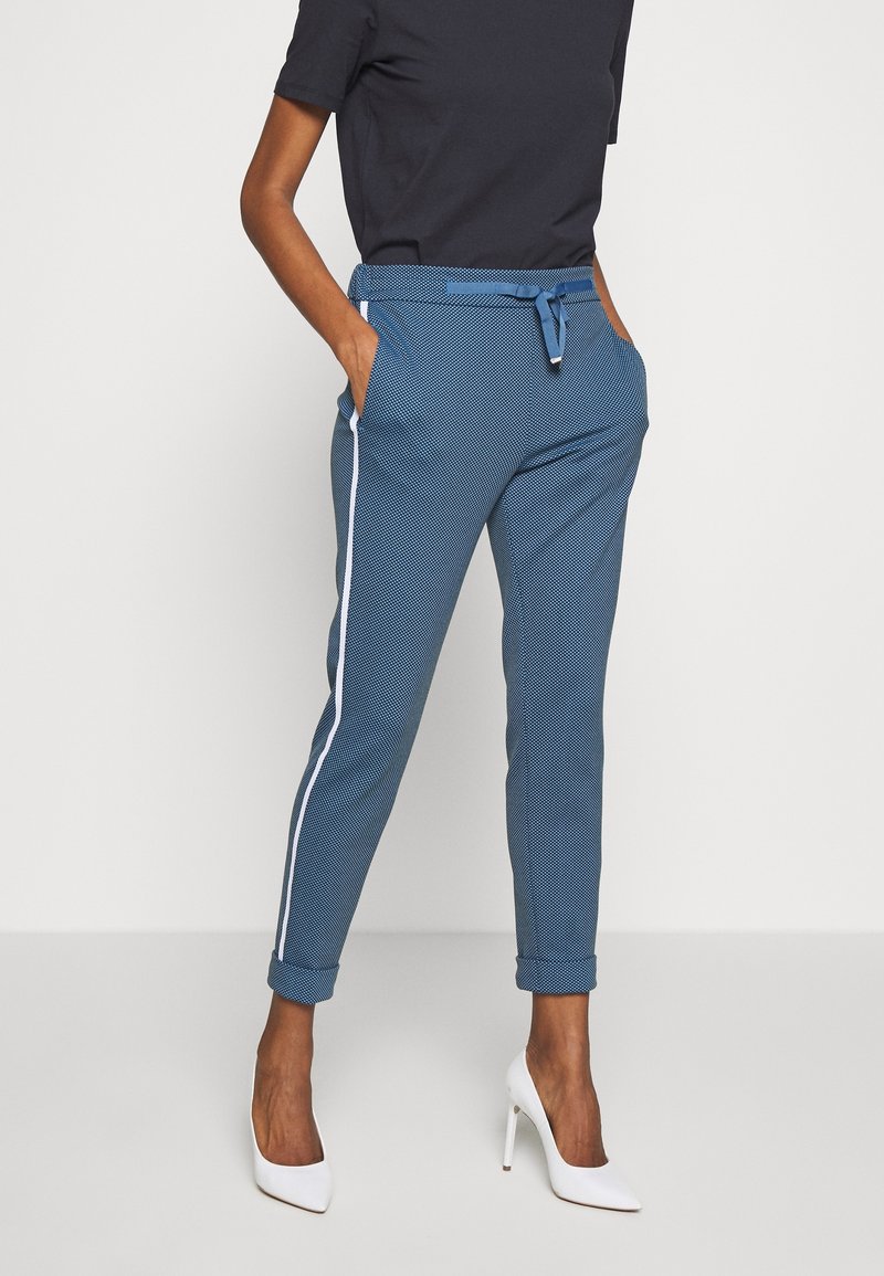 Kobieta ubrana w niebieskie, fakturowane spodnie typu tapered z białymi bocznymi paskami, mankietami i dopasowanym paskiem, w połączeniu z białymi butami na wysokim obcasie.