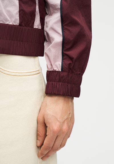 Close-up af en hånd, der hviler ved siden af en cremefarvet buks' lomme, iført en mørkerød og lyserød langærmet jakke med elastiske manchetter.