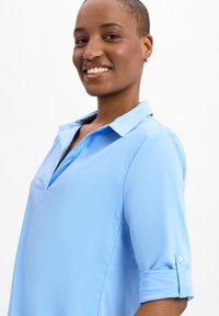 Camicia azzurro chiaro con colletto, chiusura con bottoni, maniche corte arrotolate, taschino sul petto e una texture liscia.