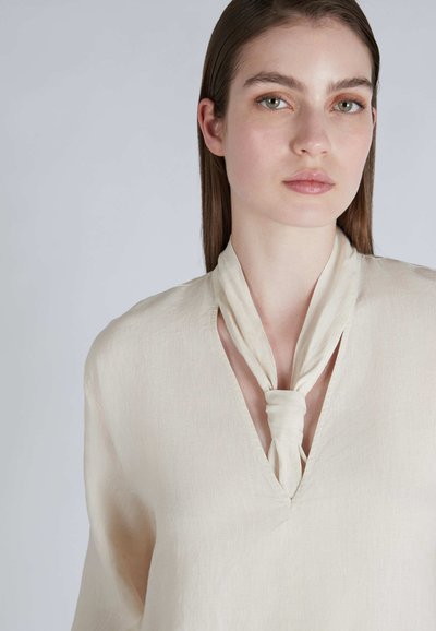 Cremefarbene, leichte Bluse mit V-Ausschnitt und einem gebundenen Stoffdetail am Kragen. Weiche Textur, lange Ärmel und lockere Passform.