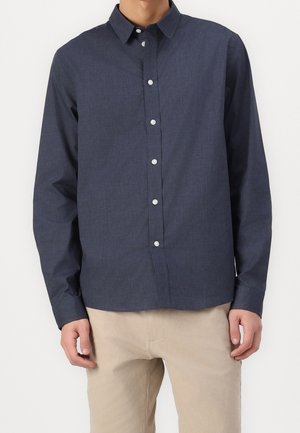 Mann trägt ein dunkelblaues Hemd mit langen Ärmeln und helle beige Hose, steht vor einem weißen Hintergrund.