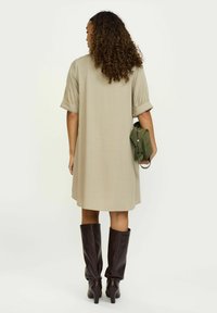 Bēša ceļgalu garuma kleita ar īsām, pūkainām piedurknēm un teksturētu apdari, apvienota ar tumši brūnām ceļgalu augstām zābakiem un zaļu somu.