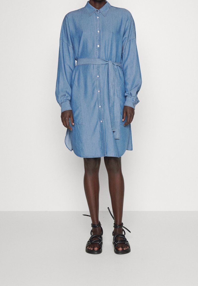 Personne portant une robe en jean bleu clair boutonnée avec manches longues et ceinture à la taille, associée à des sandales plates noires à brides.