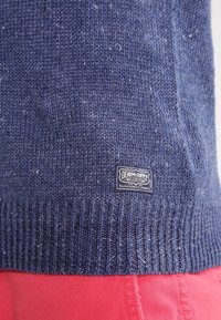Maglione a maglia blu navy con orlo a costine; presenta un'etichetta nera con la scritta "DENIM&SUPPLY". Il primo piano mostra la texture e una leggera granatura.