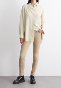 Camicia a maniche lunghe beige chiaro con colletto, abbinata a pantaloni beige aderenti e scarpe chunky nere con lacci. Tessuto di trama liscia.