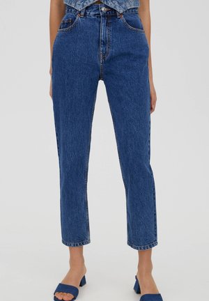 Blaue Jeans mit hoher Taille und geradem Bein, getragen mit blauen Sandalen mit Absatz und offener Spitze, von der Taille bis zu den Knöcheln vor einfarbigem Hintergrund gezeigt.