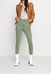 Chaqueta de cuero marrón sobre una blusa floral, combinada con jeans skinny verdes y botas blancas hasta el tobillo. El conjunto presenta una silueta ajustada.