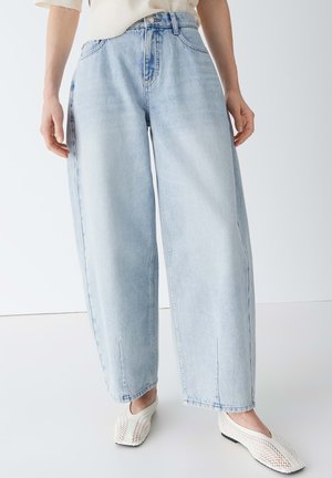 Person trägt hellblaue Jeans mit weitem Bein und weiße, gewebte Ballerinas, steht vor einem schlichten weißen Hintergrund.