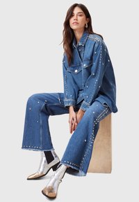 Conjunto de chaqueta y pantalones de mezclilla con acentos de tachuelas plateadas; la chaqueta presenta dos bolsillos en el pecho, un corte holgado y un dobladillo desflecado; botas metálicas brillantes.