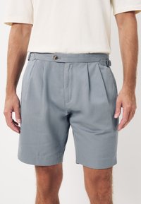 Pantaloncini grigi realizzati in tessuto morbido, caratterizzati da un design plissettato, chiusura con bottoni e cinturini laterali regolabili per una vestibilità su misura.