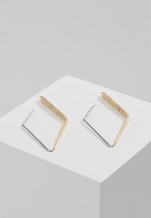 Paire de boucles d’oreilles géométriques avec un design angulaire en métal doré et argenté, posées sur un piédestal blanc en forme de cube.