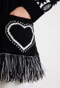 Black knitted cardigan featuring a large white heart design on the pocket, detailed with fringe at the hem in black and white.