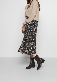 Czarna plisowana spódnica midi w kwiaty z różowymi i zielonymi wzorami, zestawiona z beżowym dzianinowym swetrem i brązowymi botkami za kostkę. Torebka w kolorze taupe.