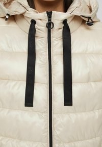 Chaqueta tipo puffer en beige claro con acabado brillante, presenta un zipper negro y cordones, además de una capucha para mayor abrigosidad.