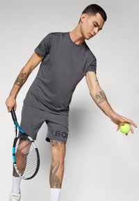Graues Sport-T-Shirt und Shorts mit Logo-Akzenten. Glatte Textur. Hält einen Tennisball und einen Schläger. Tätowierungen sind am Arm sichtbar.