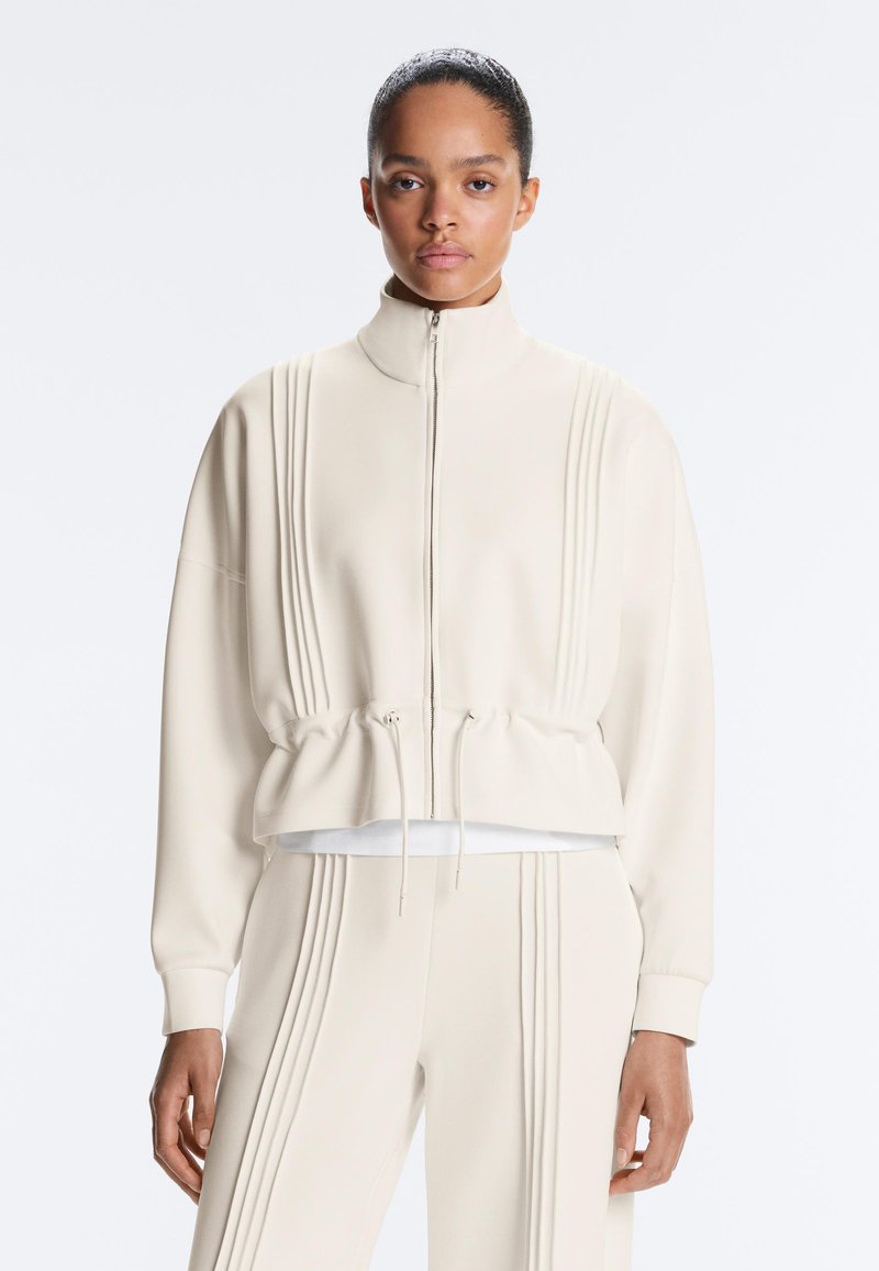 Jeune femme portant une veste crème zippée et un pantalon assorti avec des détails de coutures verticales, debout devant un fond uni.